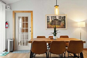 Ein Essbereich mit Holztisch, vier Stühlen, einer Topfpflanze, einem gerahmten Bild an der Wand, einem Feuerlöscher und einem Mülleimer.