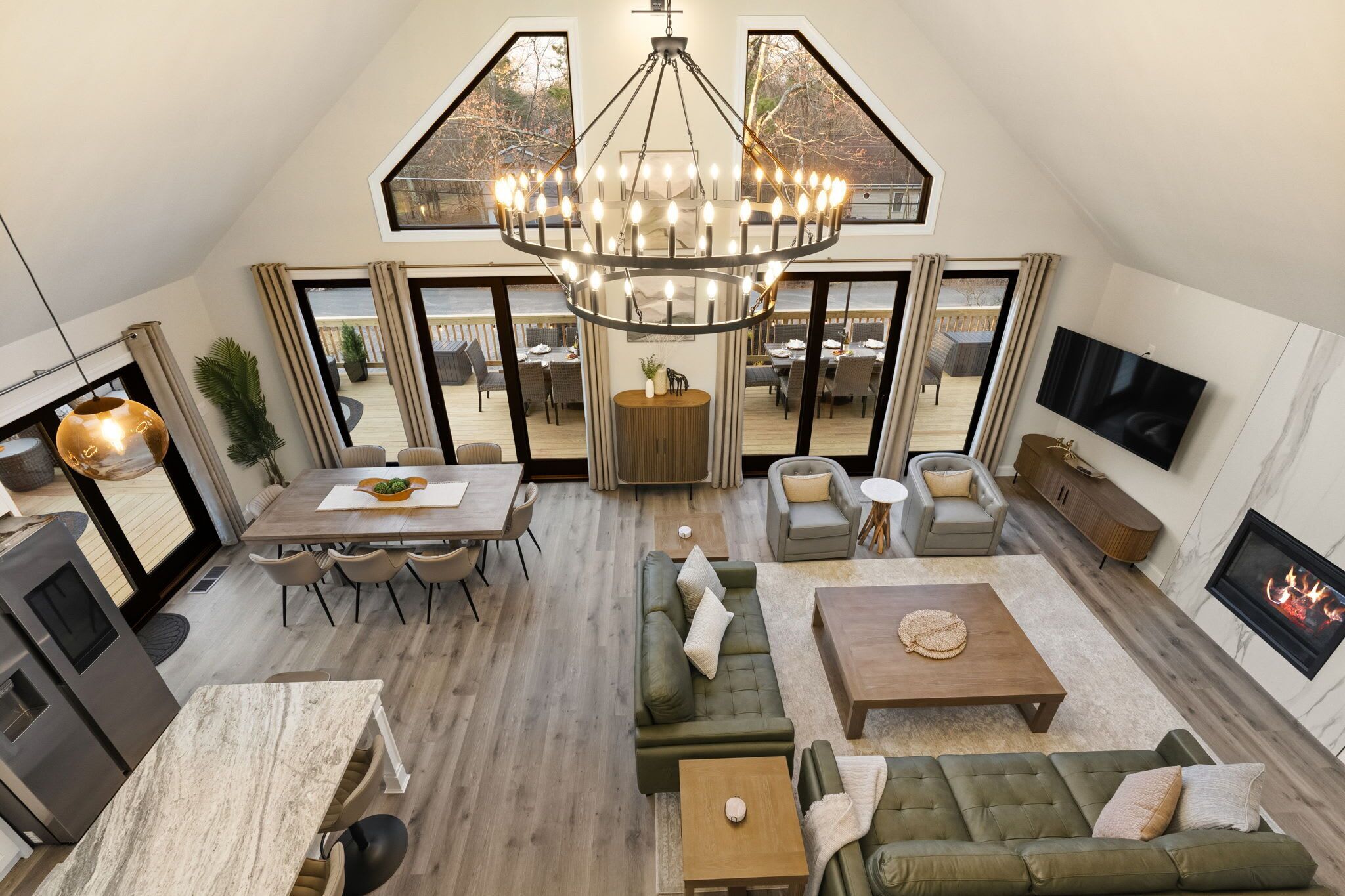 A spacious loft-style living area with a large chandelier, a dining table, sofas, a fireplace, and large windows offering views of the outdoors.