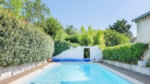 Una piscina con cubierta azul, rodeada por un muro blanco y abundante vegetación.