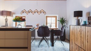A modern kitchen with a central island, wooden cabinets, a dining area with chairs, and a hanging light fixture. - Klochberg R&L APP Sunrise - Gelber Muskateller (Kloech)
