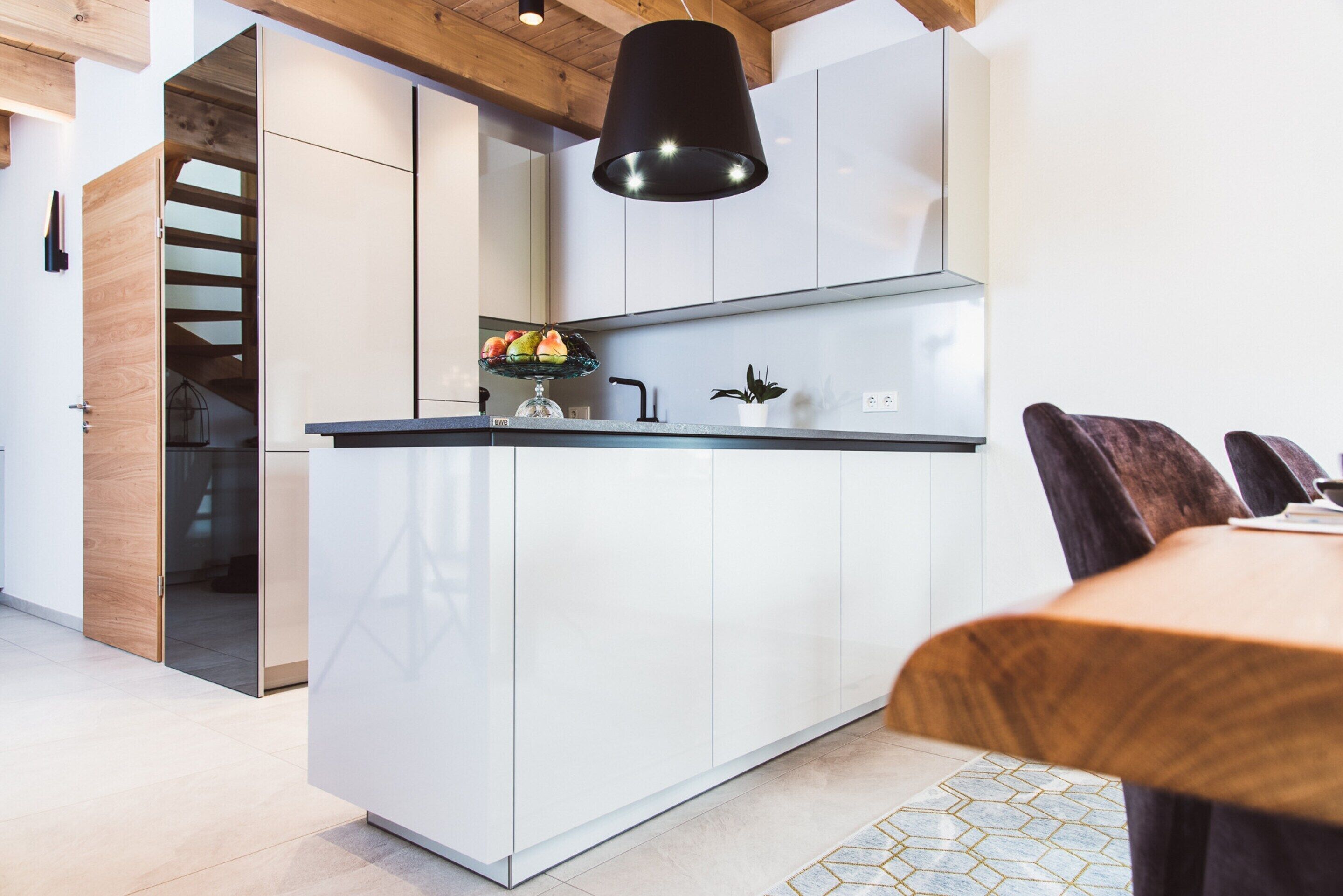 Una cocina moderna con armarios blancos, una lámpara colgante negra y una zona de comedor con mesa y sillas de madera.