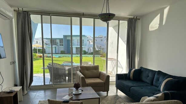 Ein Wohnzimmer mit Sofa, Sessel und Couchtisch, große Fenster mit Blick auf einen Pool und Gebäude.