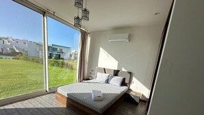 Ein modernes Schlafzimmer mit einem großen Bett, Blick auf eine Grünfläche und einem Fenster mit Vorhang.