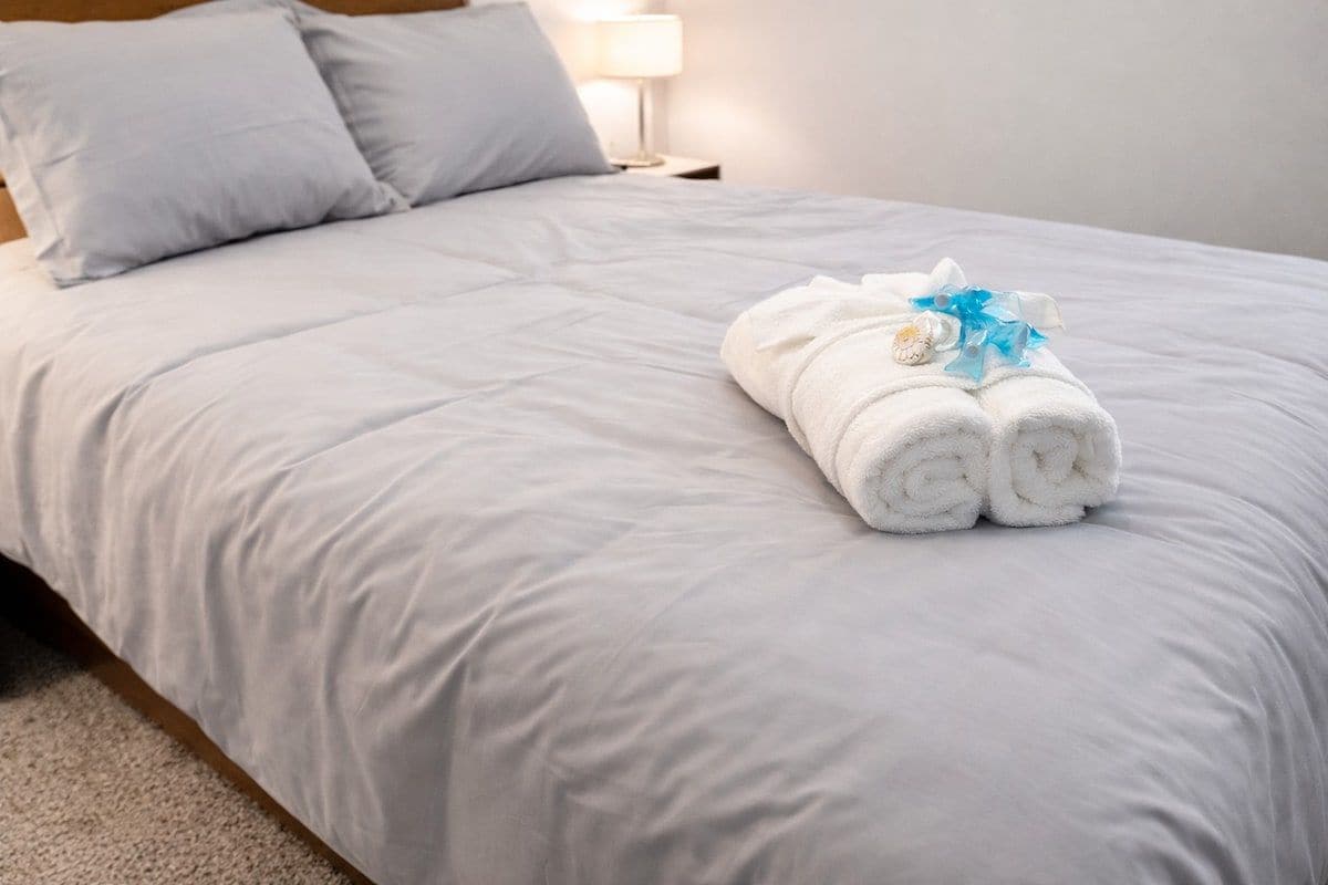 A neatly made bed with a white comforter and pillows, a rolled white towel with a blue ribbon, and a small decorative item on the bed.