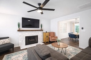 A living room with a fireplace, a television, a ceiling fan, a sofa, a coffee table, and a dining area with chairs.