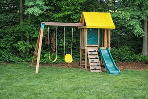 A wooden swing set with a yellow roof, a slide, and a climbing ladder, surrounded by a grassy area and trees.