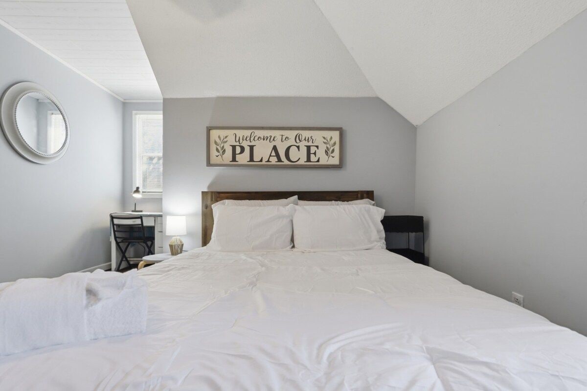 Una cama bien hecha en una habitación con un cartel en la pared que dice 'Welcome to Our PLACE', un espejo redondo y una mesita con una lámpara.