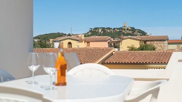 Une terrasse avec des chaises blanches et une table, d’où l’on peut admirer une ville aux bâtiments aux toits de tuiles et une colline surmontée d’une tour.