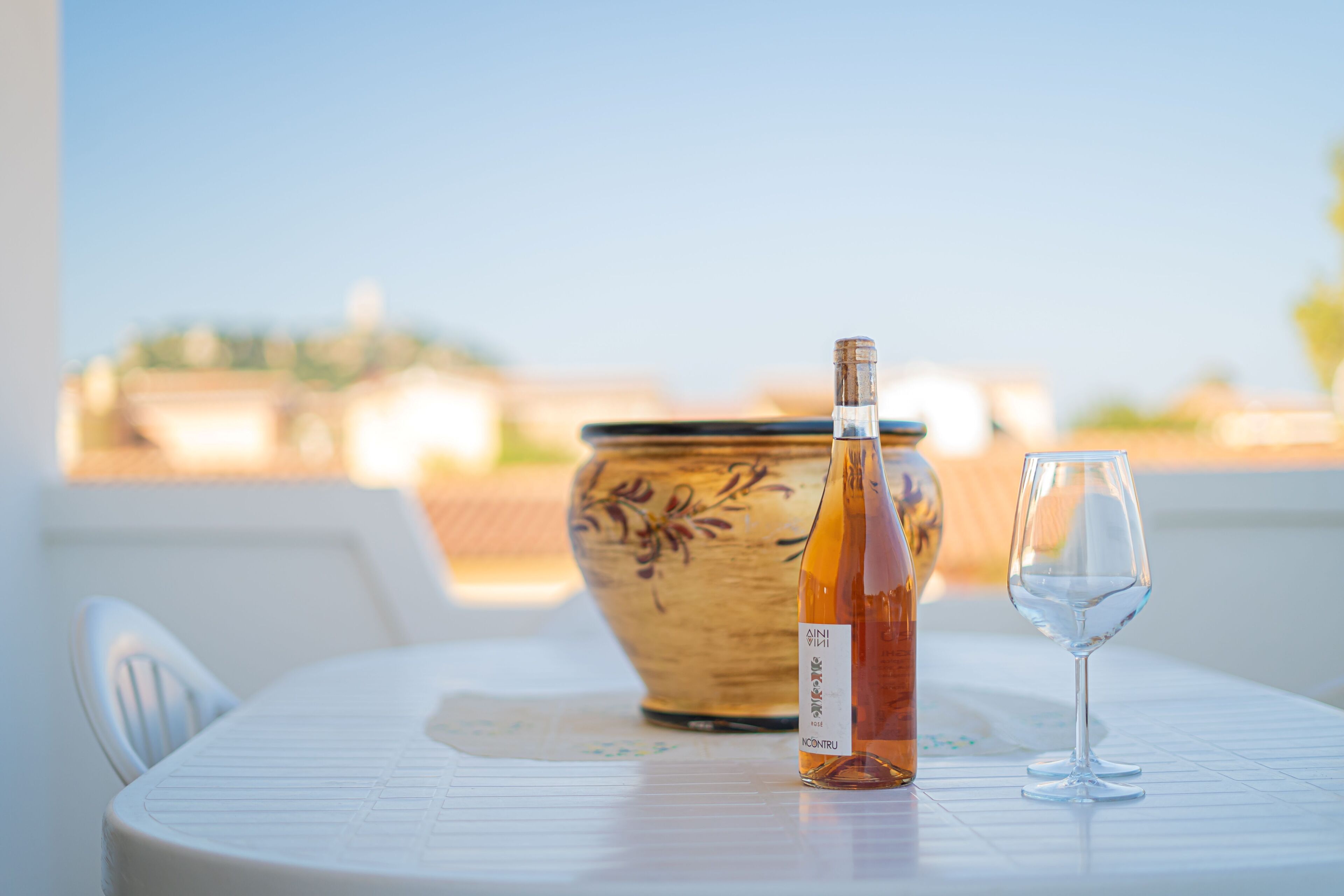 A table with a bottle and a glass on a white tablecloth, a decorative bowl, and a view of buildings in the background.