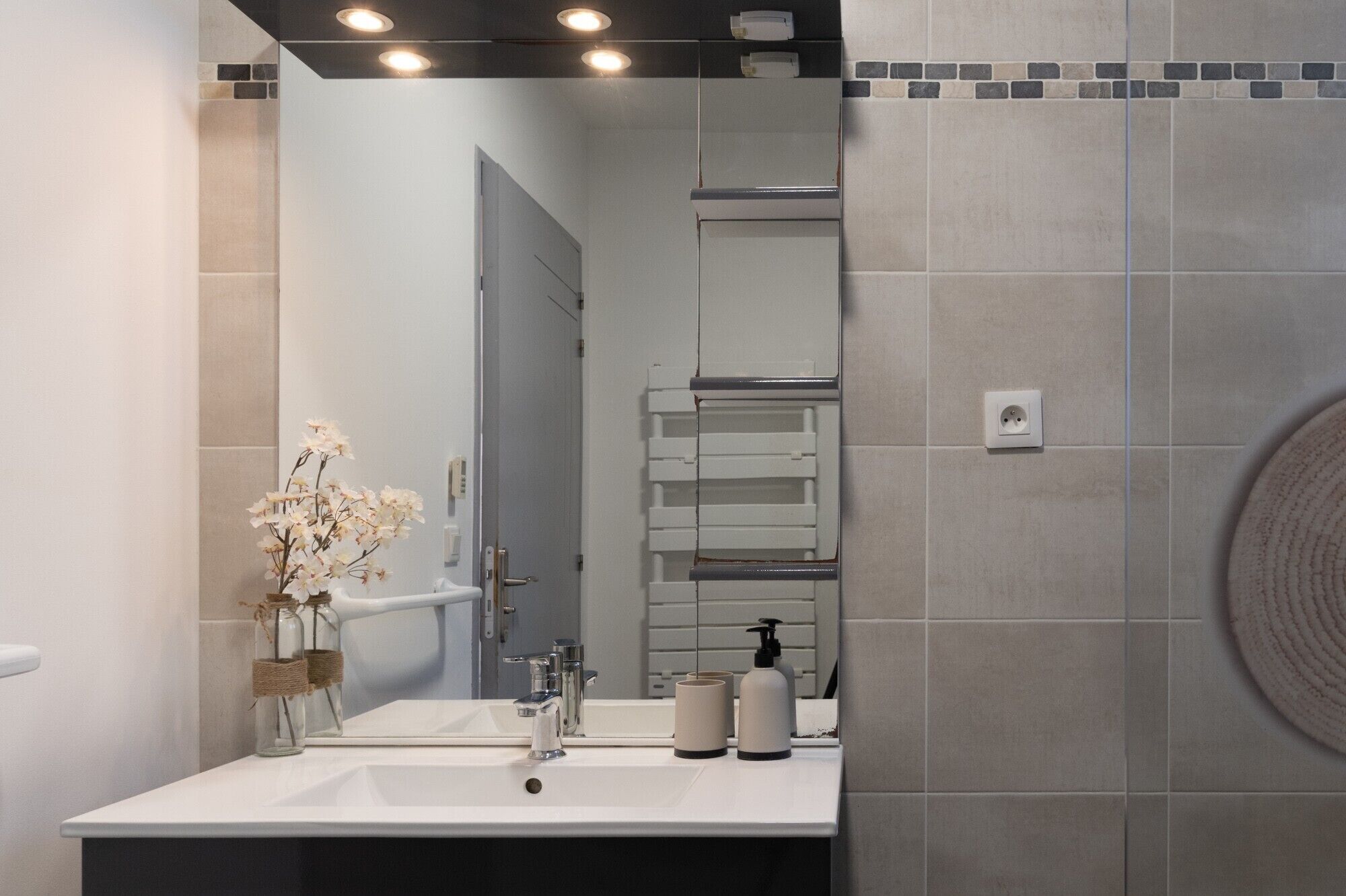 Un baño con lavamanos blanco, dos espejos, toallero, dispensador de jabón y zona de ducha.