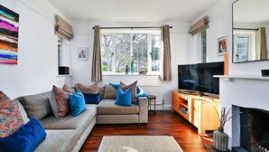 Ett vardagsrum med en soffa, ett tv-appar, en öppen spis och stora fönster.