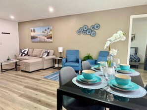 Ein Wohnzimmer mit einer Couch, Sesseln, einem Esstisch gedeckt mit blauen Tellern und Gläsern, einem Bild von einer Strandlandschaft und einer Wanddekoration aus runden Objekten.