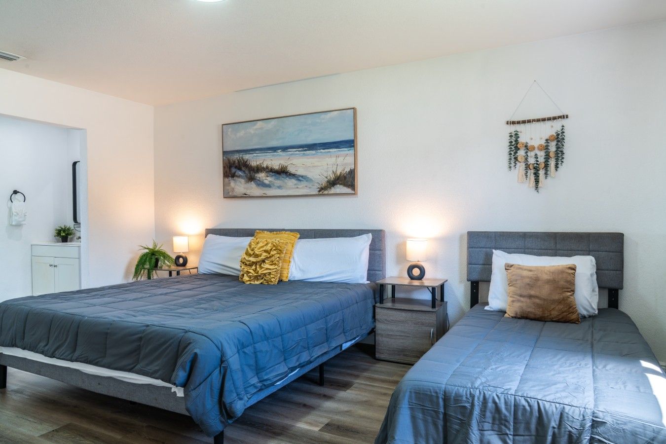 Bedroom: Unwind in comfort in this bright guest bedroom featuring a spacious queen bed and an additional twin bed, adorned with soft neutral linens, warm bedside reading lights, a classic dress display, and coastal-inspired wall art.