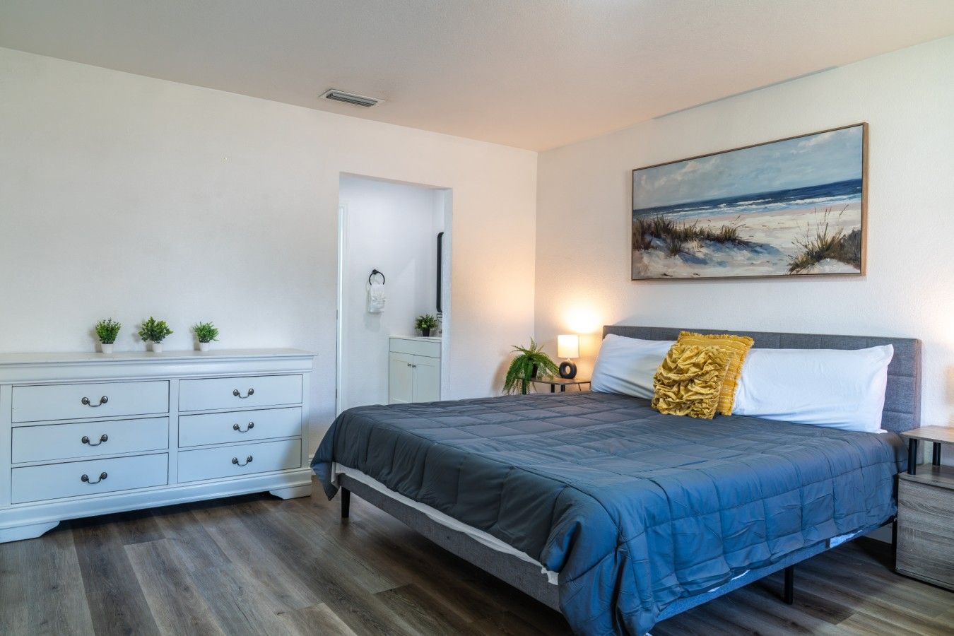 Spacious Dual-Bed Retreat: Rest and recharge in this airy bedroom complete with a plush queen mattress, a large white storage dresser with ample drawer space, serene beach artwork, and warm ambient lighting.