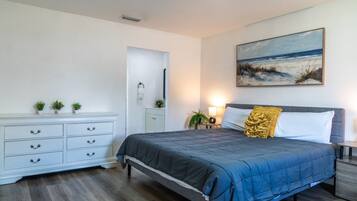 Spacious Dual-Bed Retreat: Rest and recharge in this airy bedroom complete with a plush queen mattress, a large white storage dresser with ample drawer space, serene beach artwork, and warm ambient lighting.