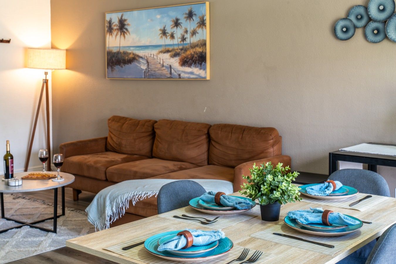 Open-Concept Living & Dining Area: Unwind and entertain with ease in this spacious open-concept living space, featuring a plush sectional sofa, warm tripod floor lamp, dedicated dining setup, and coastal artwork that sets a relaxed beachy tone.