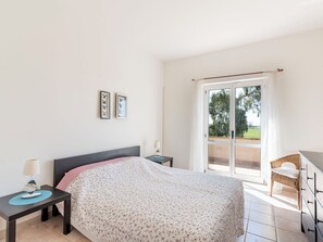 Ein Schlafzimmer mit einem Bett, einem Nachttisch, einem Sessel, einer Kommode und einem Fenster mit Blick auf Grünflächen.