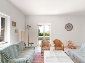 Ein großzügiges Wohnzimmer mit einer Couch, zwei Korbstühlen, einem Glastisch und Blick durch eine Glastür.