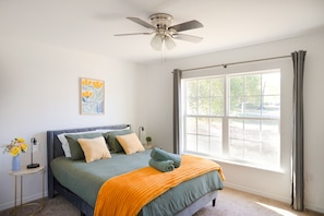 A bedroom with a bed, a ceiling fan, a window with curtains, and a wall-mounted artwork.