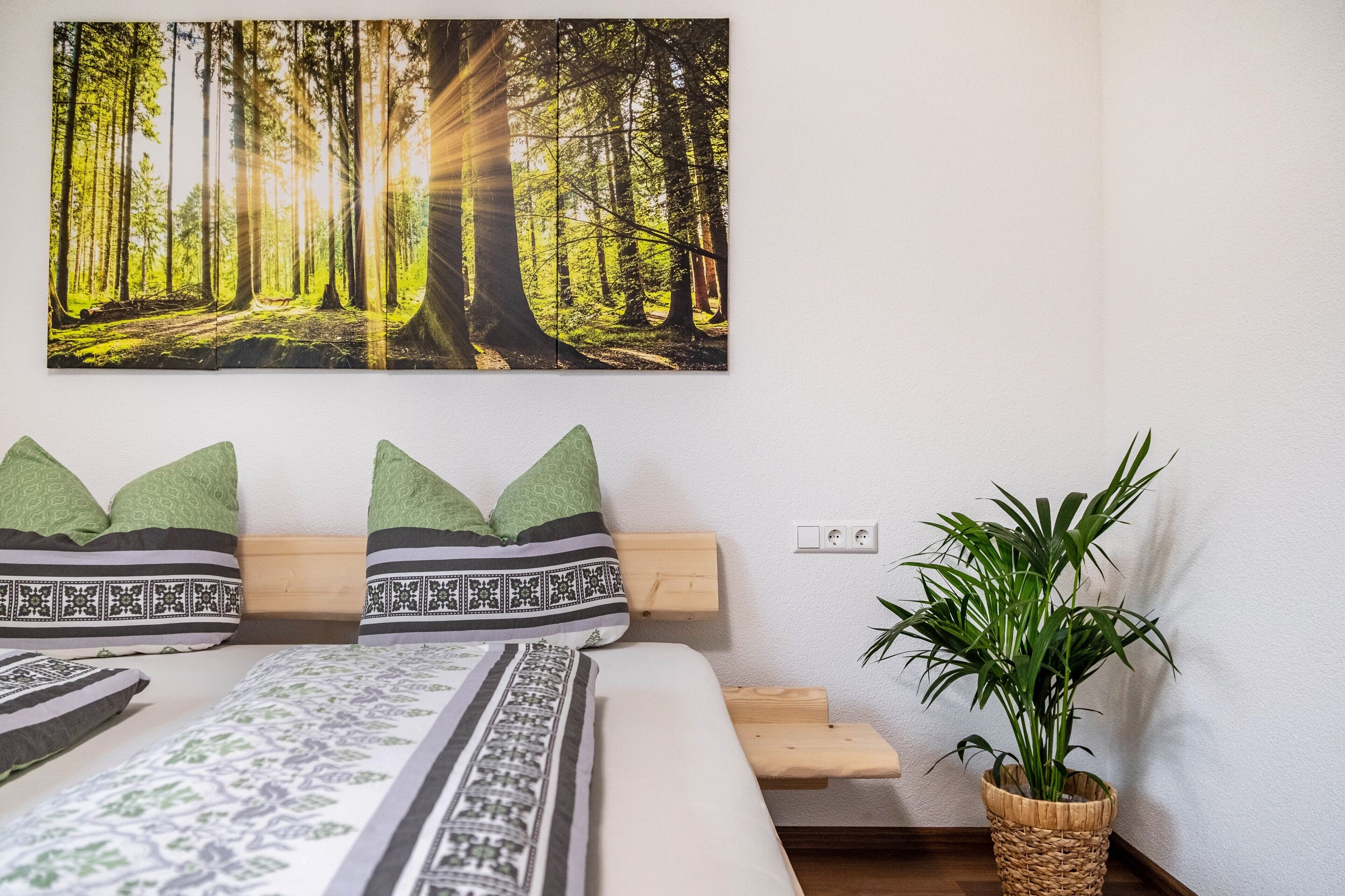 A bedroom with a bed, pillows, a bedside table, a wall-mounted painting of a forest, and a potted plant.