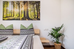 Une chambre à coucher avec un lit, des oreillers, une table de chevet, un tableau représentant une forêt accroché au mur et une plante en pot.