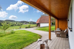 Une terrasse couverte meublée de mobilier en osier, avec vue sur un paysage rural, et un bâtiment doté d’un toit recouvert de tuiles rouges.