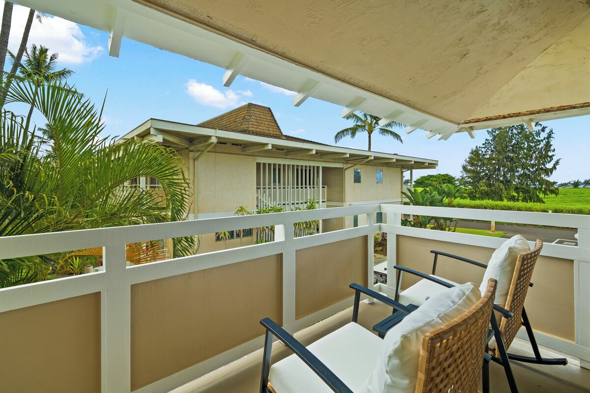 A balcony with a white railing, two wicker chairs with cushions, and a view of a house and greenery.