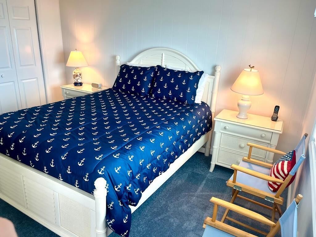A bed with a navy blue bedspread, two bedside tables with lamps, a wooden chair with an American flag pillow, and a white door in the background.