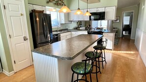 A kitchen with a central island, bar stools, pendant lighting, and a refrigerator. - Beachside Condo / Linens Provided! By Williamson Realty (Ocean Isle Beach)