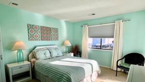 A bedroom with a bed, two bedside tables, a window with curtains, a chair, and a wall with patterned artwork. - Beachside Condo / Linens Provided! By Williamson Realty (Ocean Isle Beach)