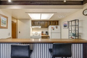 Eine Küche mit Holzausstattung, einem weißen Kühlschrank, einer Kaffeemaschine und einem Metallregal. Vor der Arbeitsplatte stehen zwei schwarze Stühle.
