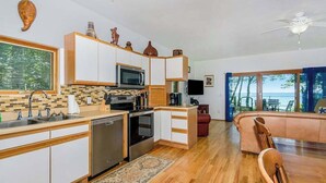 A modern kitchen with a microwave, stove, and dishwasher, a dining area with a wooden table and chairs, and a view of the ocean through large windows. - Dog-friendly home with lake views, wood stove, kayaks/SUPs, & W/D (Coloma)