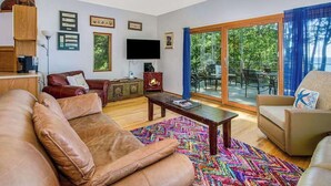A living room with a sofa, armchair, coffee table, and a TV. There is a large window with a view of trees. - Dog-friendly home with lake views, wood stove, kayaks/SUPs, & W/D (Coloma)