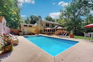 Ein rechteckiger Swimmingpool mit Liegestühlen und Sonnenschirmen, umgeben von einem Haus und Bäumen.