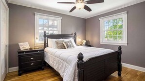 Une chambre à coucher avec un lit, des tables de chevet, un ventilateur de plafond et deux fenêtres.