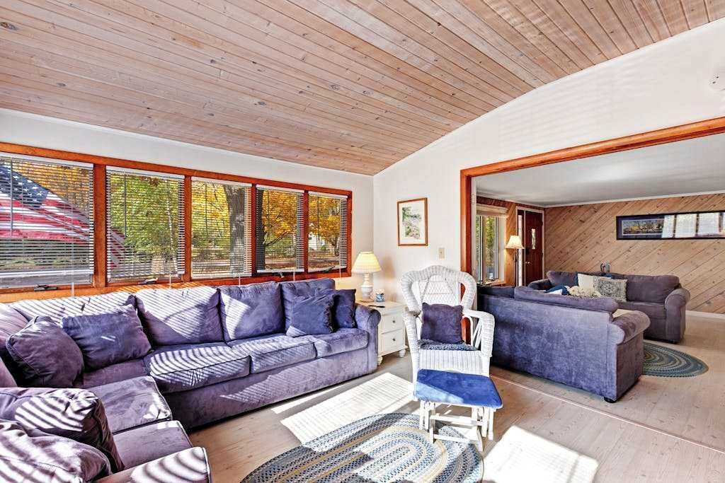 Amplia sala de estar con un sofá grande, una silla y una mesita. Hay ventanas grandes con persianas, techo de madera y vista a árboles.
