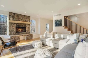 Una sala de estar con chimenea de piedra, un sofá gris, una silla de madera y una escalera.