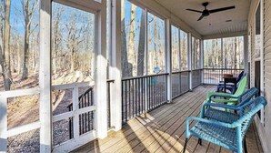 A covered porch with a ceiling fan, wooden railing, and a view of a forest.