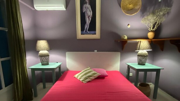 A bedroom with a red bedspread, two bedside tables with lamps, a wall-mounted air conditioner, a framed artwork, and a decorative vase. - Villa-Agave, by the sea in Porto-Rafti outside ATH (Porto Rafti)