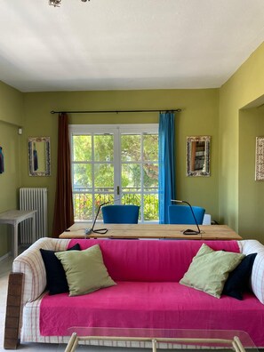 Ein Zimmer mit großem Fenster, einem Tisch mit Stühlen, einer Couch mit Kissen und einem Heizkörper.