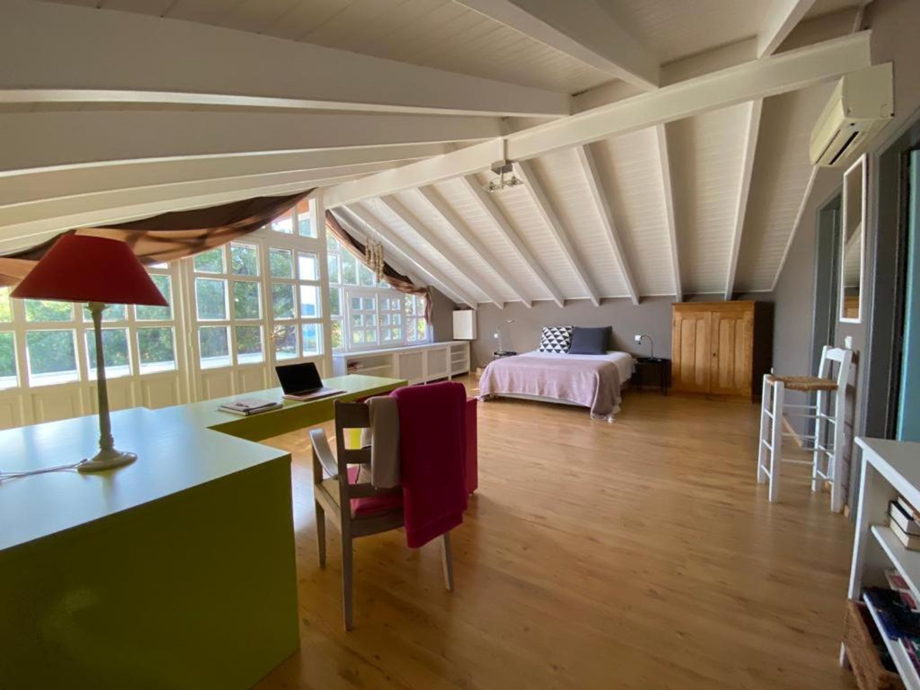 A spacious attic bedroom with a wooden floor, a desk with a laptop, a bed with a pink blanket, and a window with curtains.