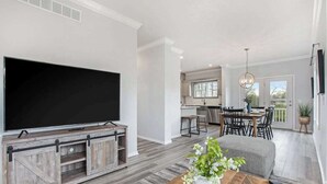 Un salon moderne doté d’un grand téléviseur à écran plat, d’un meuble de rangement en bois, d’un coin repas avec une table et des chaises, et d’une cuisine en arrière-plan.