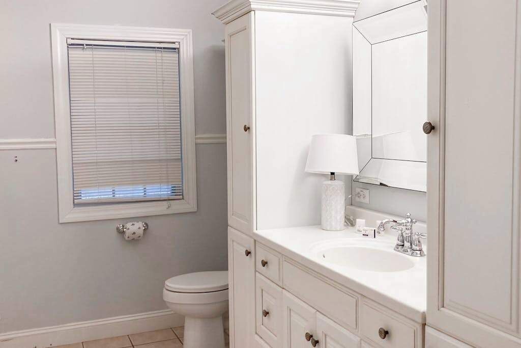 Une salle de bain comprenant des toilettes, un lavabo avec un miroir et une fenêtre avec des stores.