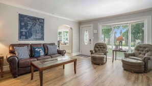 Sala de estar con un sofá de cuero marrón, una mesa de centro de madera y dos sillones. Hay un mapa en la pared y una puerta que da a un balcón.
