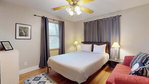 Ein Schlafzimmer mit einem Bett, einem Deckenventilator, einem Nachttisch, einer Lampe, einem Sofa, einem Teppich und einem Fenster mit Vorhängen.
