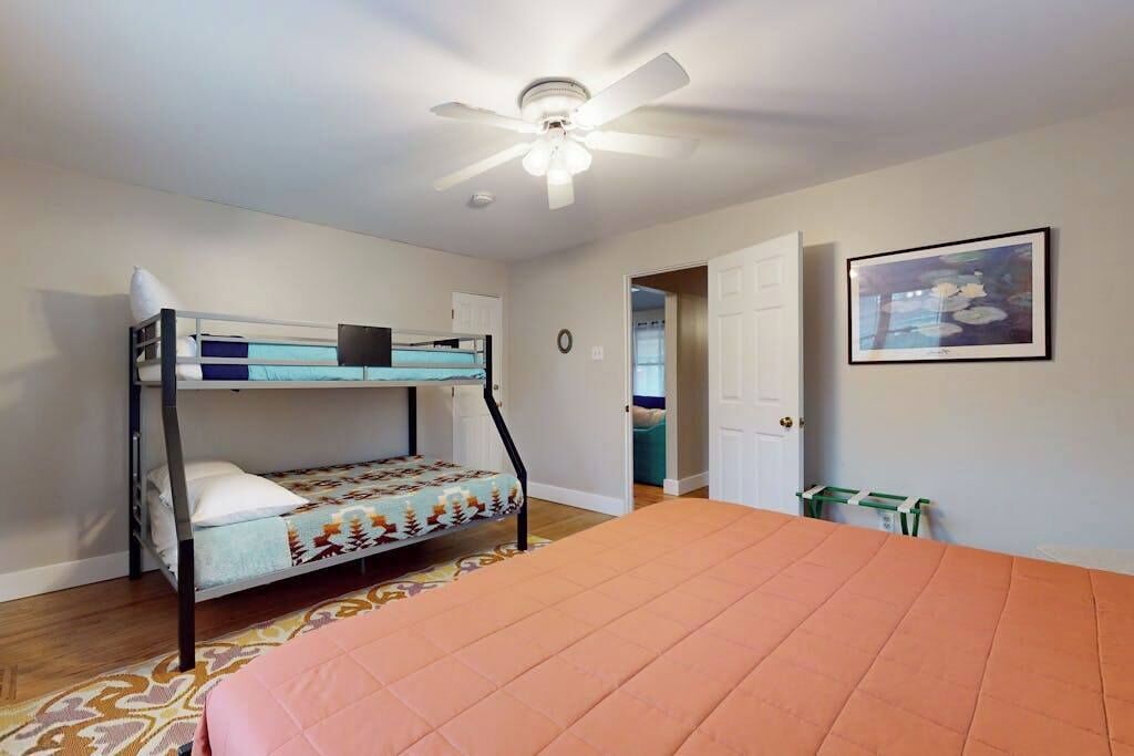 A room with bunk beds, a ceiling fan, a bed with an orange bedspread, a framed picture on the wall, and a door leading to another room.