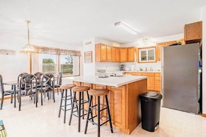 A kitchen with wooden cabinets, a central island with stools, a refrigerator, and a dining area with chairs.