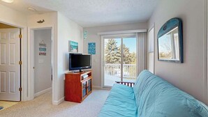 A living room with a TV, a blue couch, and a balcony with a view of trees. - Charming 3 story condo, w/ a W/D & Wifi, near beaches & shared pool access (Manistee)