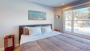 A bedroom with a bed, a painting of a lighthouse, a window with a view, and a small bedside table. - Charming 3 story condo, w/ a W/D & Wifi, near beaches & shared pool access (Manistee)