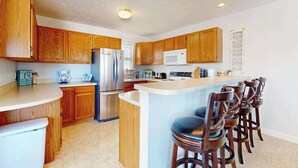 A kitchen with wooden cabinets, a stainless steel refrigerator, a microwave, and a bar area with stools. - Charming 3 story condo, w/ a W/D & Wifi, near beaches & shared pool access (Manistee)